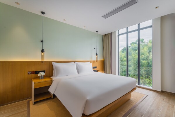Una habitación de hotel moderna con una cama grande, cabecera de madera, dos mesitas de noche con lámparas, una mesita auxiliar con un jarrón de flores y un ventanal con vistas a zonas verdes.