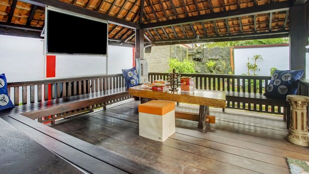 A covered outdoor seating area with wooden benches, a table with a cushion, and a TV mounted on the wall. - OYO Life 3982 Azka Homestay Syariah (Bandar Lampung)