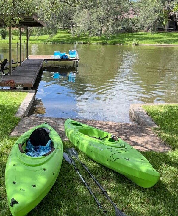 A grassy area with two kayaks and a paddle, a dock with chairs, and a calm pond.