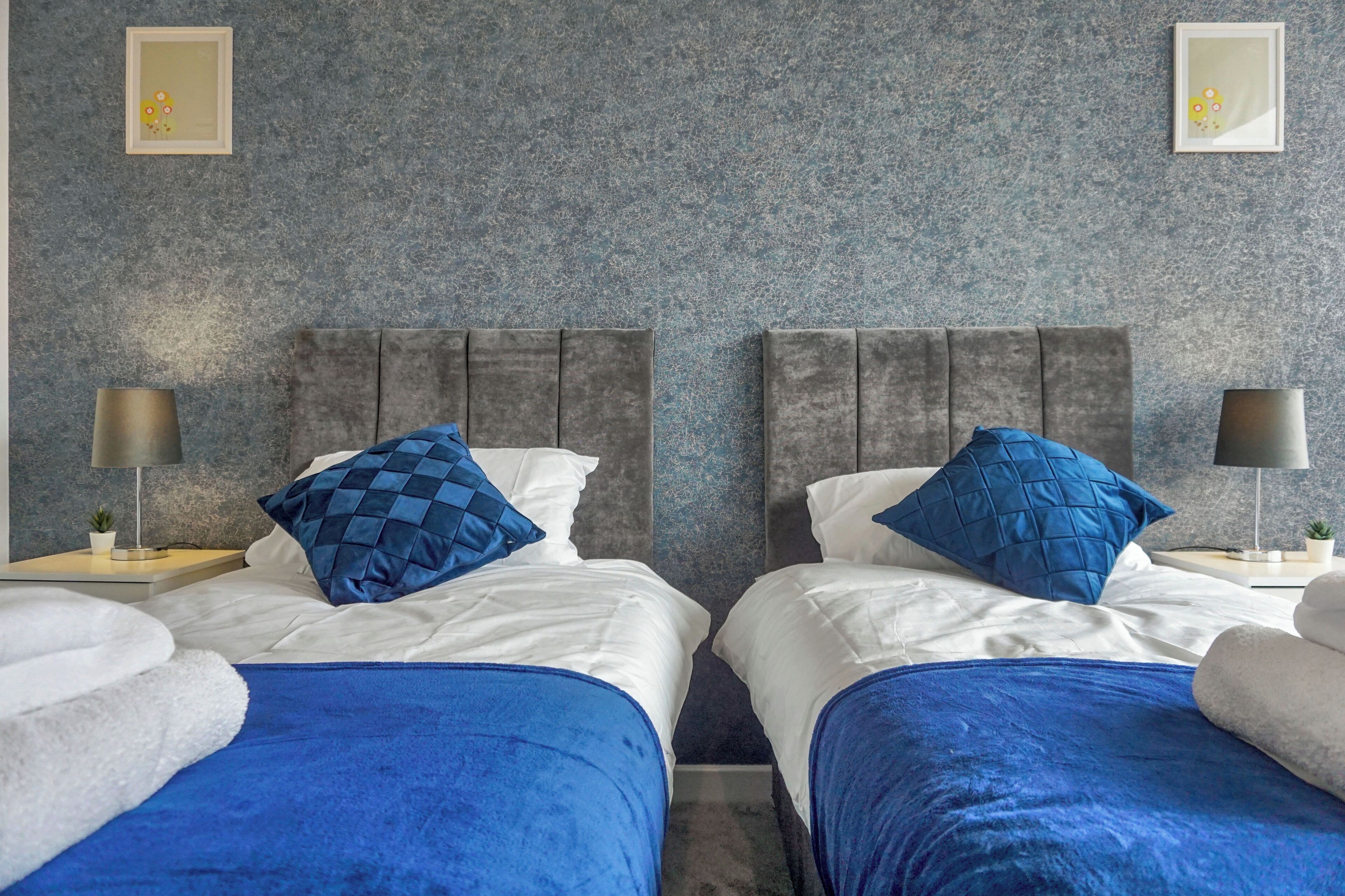A hotel room with two beds, each with a blue blanket and a blue quilted pillow. There are two bedside tables with lamps and small plants. The walls are textured and there are framed pictures on the wall.