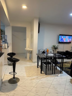 Una sala de estar moderna con comedor, televisor montado en la pared y una cocina visible al fondo.