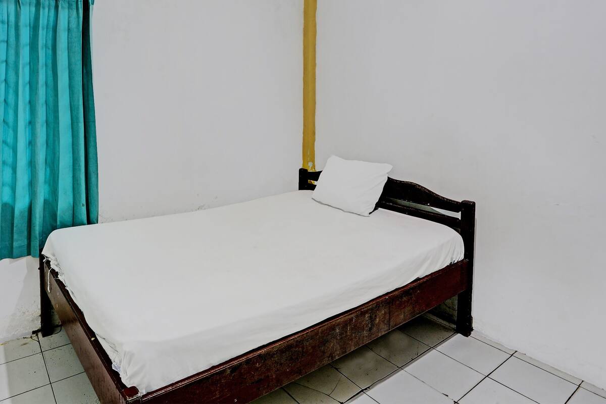 Un lit simple avec un matelas blanc et un cadre en bois, dans une pièce aux murs blancs et au sol carrelé.