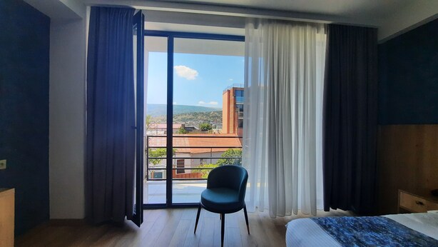 A hotel room with a large sliding glass door leading to a balcony, a single bed, a chair, and a view of buildings and hills. - Hotel DownTown Avlabari (Tbilisi)