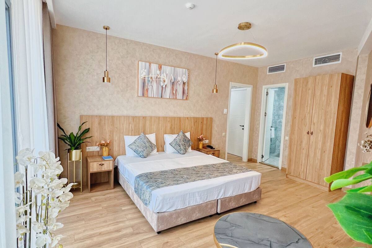 A modern bedroom with a large bed, bedside tables, a round table, and a large window.