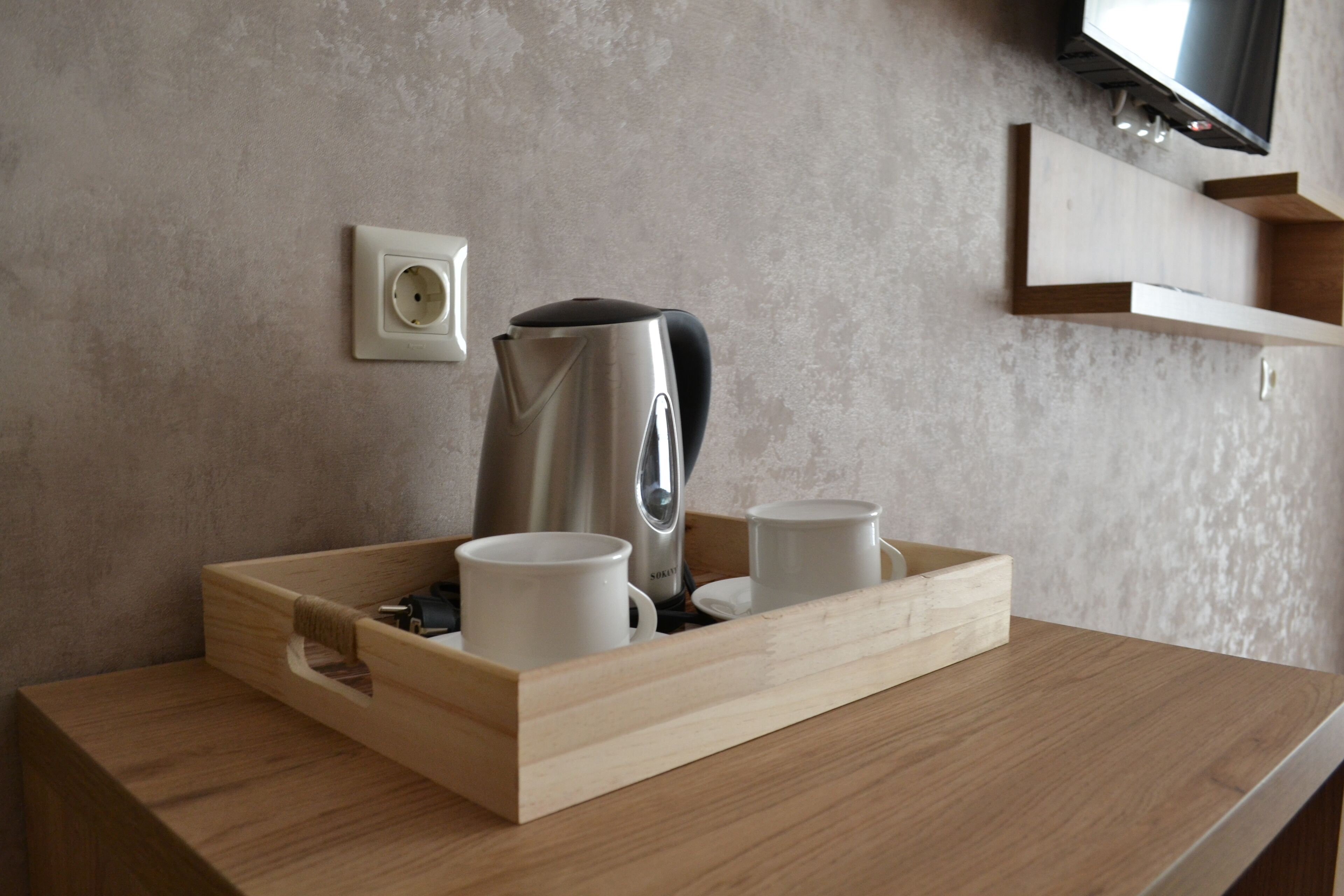 A wooden tray with two white mugs and a stainless steel coffee pot on a wooden table against a textured wall.