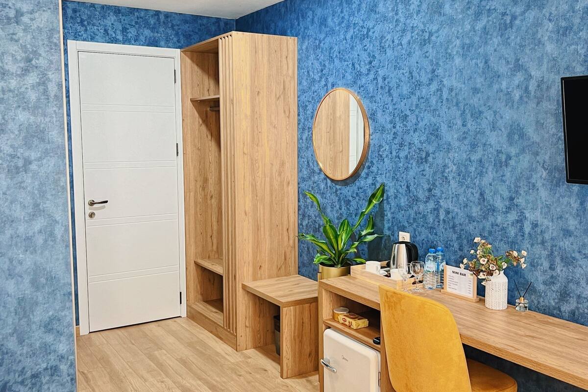 A compact room with a bed, a wooden desk, a mini fridge, a yellow chair, a round mirror, and a small potted plant.