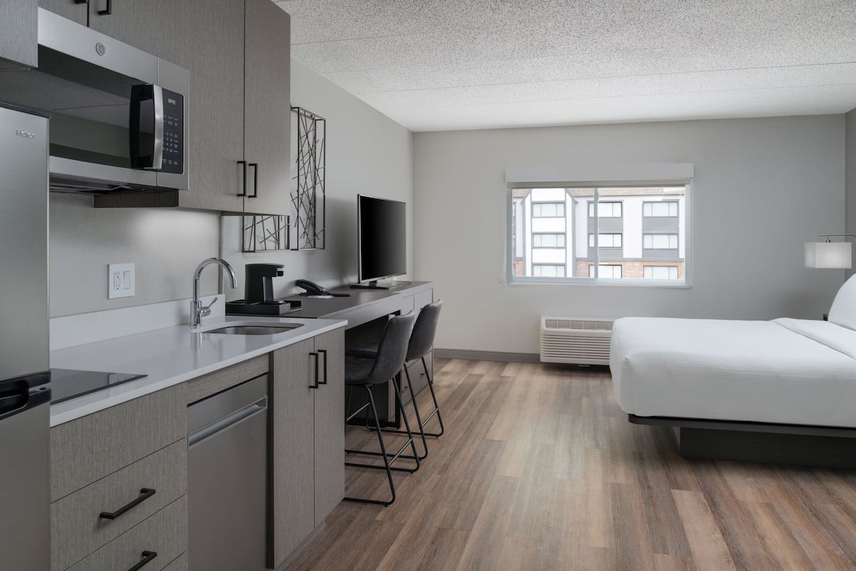 Cocina moderna con microondas, estufa y fregadero. Cuenta con una barra con sillas y un televisor de pantalla plana. También se ve una cama y una ventana con vistas a edificios.
