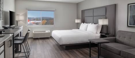 A modern hotel room with a large bed, a desk with a chair, a sofa, and a large window offering a view of the outdoors.