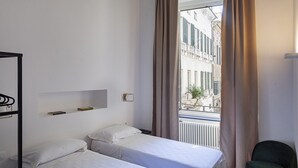 A hotel room with two single beds, a small shelf with books, a radiator, and a view of buildings through a window with curtains.