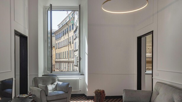 A modern living room with a sofa, armchair, and a small table with a vase. A television is on a stand, and a window offers a view of buildings.
