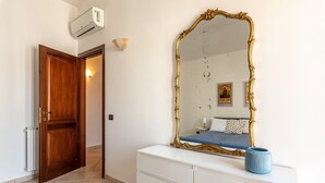 Una camera da letto con una porta in legno, una cassettiera bianca, uno specchio grande e decorato, e un vaso blu sopra la cassettiera.