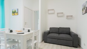 Un salon moderne avec un canapé gris, une table à manger blanche et des chaises. On y trouve trois étagères murales et une porte qui donne sur une autre pièce.