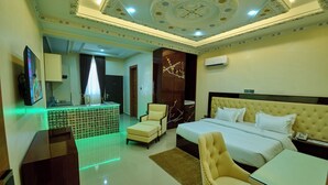 A hotel room with a large bed, a desk, a chair, and a small table. There is a TV on the wall and an air conditioning unit above the bed. - Double D Luxury Apartments (Abuja)
