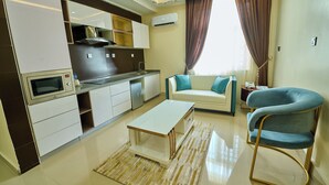 A modern kitchen with white cabinets, a microwave, and a built-in oven. There's a dining area with a white table and a blue chair. - Double D Luxury Apartments (Abuja)
