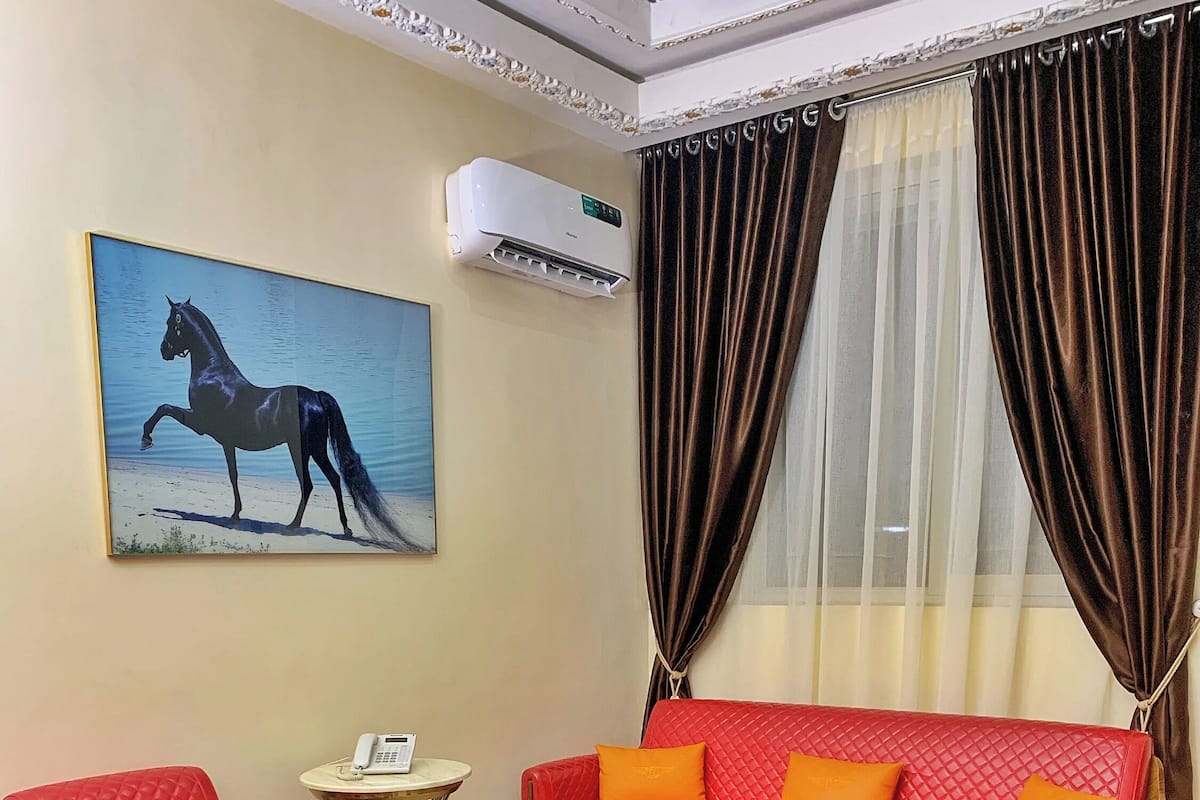A living room with a red sofa, a red armchair, and a glass coffee table. There is a painting of a horse on the wall, an air conditioner, and a window with curtains.