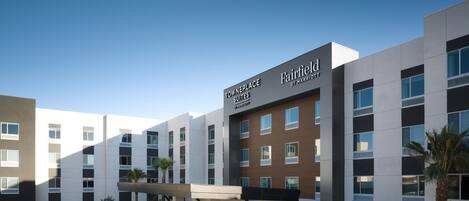 Un hotel moderno con un llamativo letrero de Fairfield by Marriott, que presenta una estructura de varios pisos con fachadas de blanco y marrón, un pasillo cubierto y una zona ajardinada con palmeras.