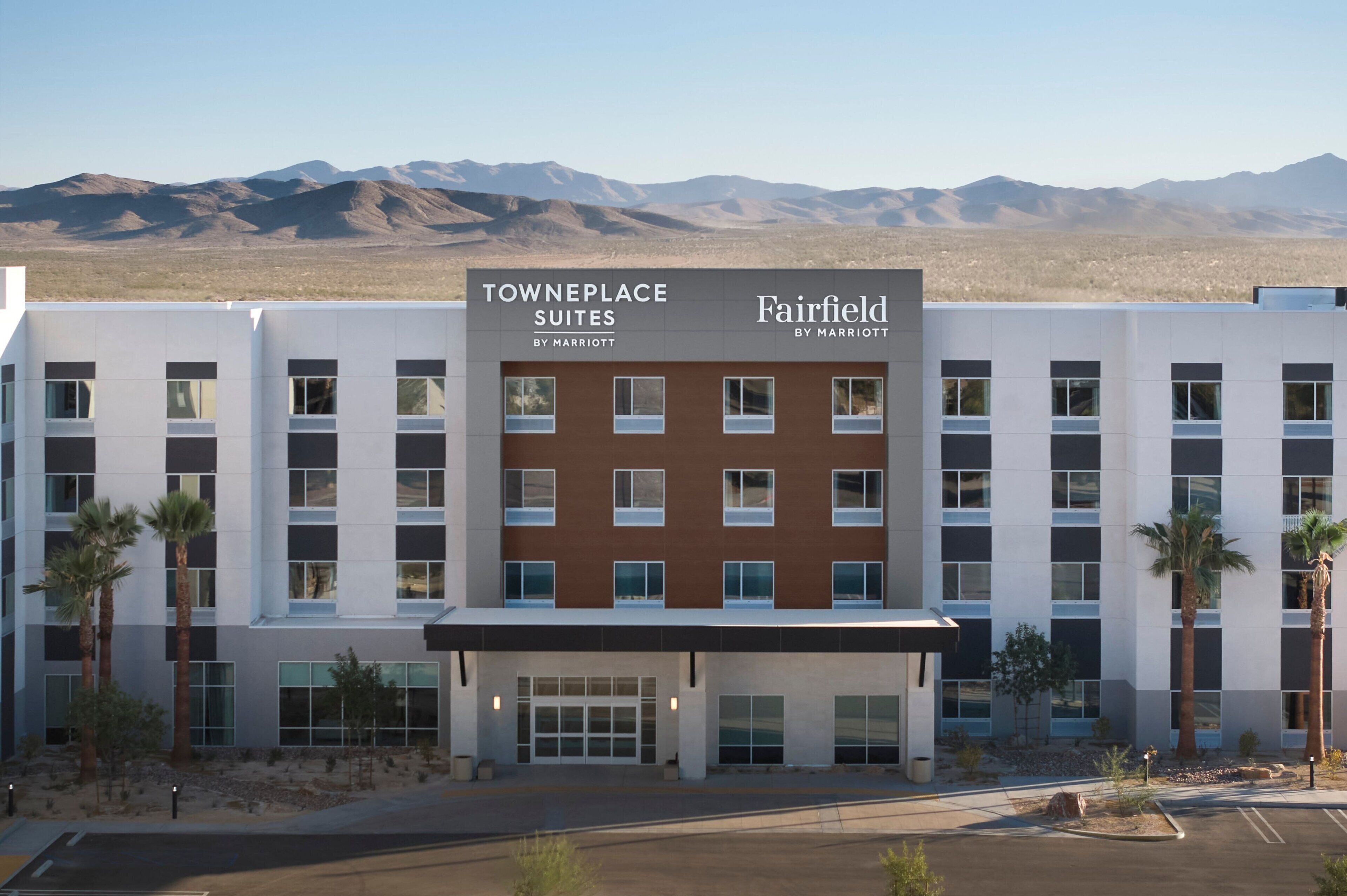 Um hotel com vários andares e design moderno, com uma combinação de painéis exteriores brancos e castanhos. O hotel chama-se "Towneplace Suites by Marriott" e tem um letreiro grande na entrada.