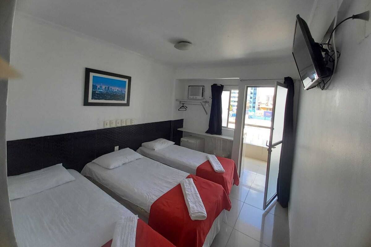 Ein Hotelzimmer mit drei Betten, einem Fernseher, einem Fenster mit Blick auf Gebäude und einem gerahmten Bild an der Wand.