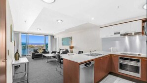 A modern kitchen with a dishwasher, oven, and stainless steel appliances. There is a dining area with a sofa and chairs, and a large window offering a view of the city. - Chevron Renaissance Private room Lvl16 (Surfers Paradise)