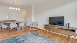 A modern living room with a wooden dining table, grey chairs, a TV on a wooden stand, and a large rug. - Barreiros Residence I (Funchal)