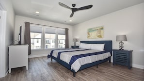 Un dormitorio con cama, escritorio, silla, ventilador de techo, ventana y un cuadro en la pared.