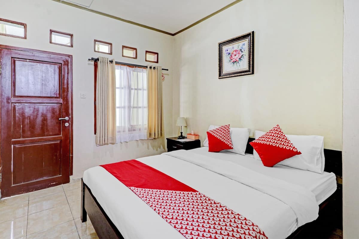 A hotel room with a large bed, a wooden door, a window with curtains, a nightstand, and a framed picture on the wall.
