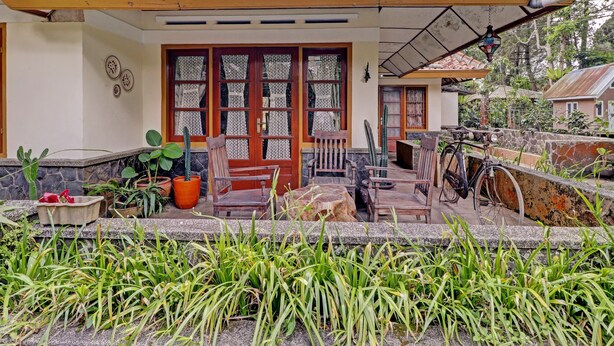 A house with a tiled roof, a porch with wooden furniture, and a garden area with plants and a cactus. - Collection O 90461 Graha Atmadja Syariah Guest House (Lembang)