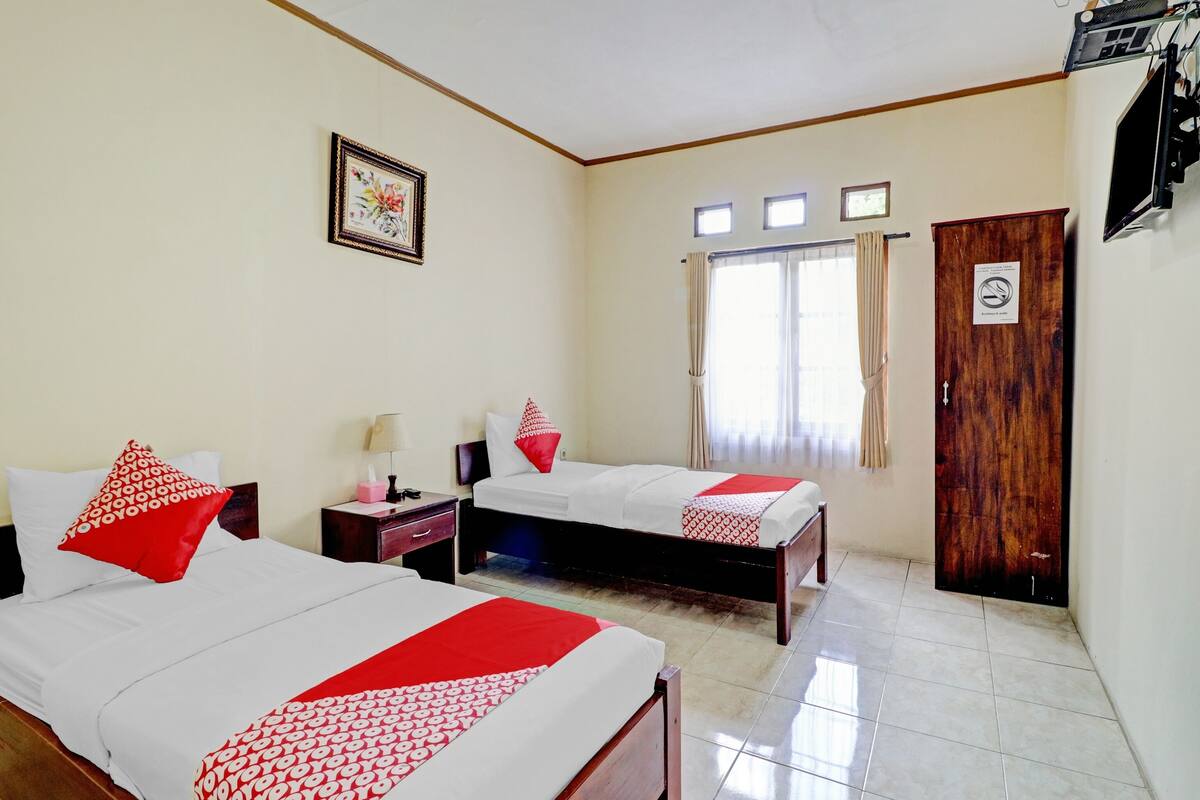 A hotel room with two beds, a wooden wardrobe, a framed picture on the wall, and a window with curtains.