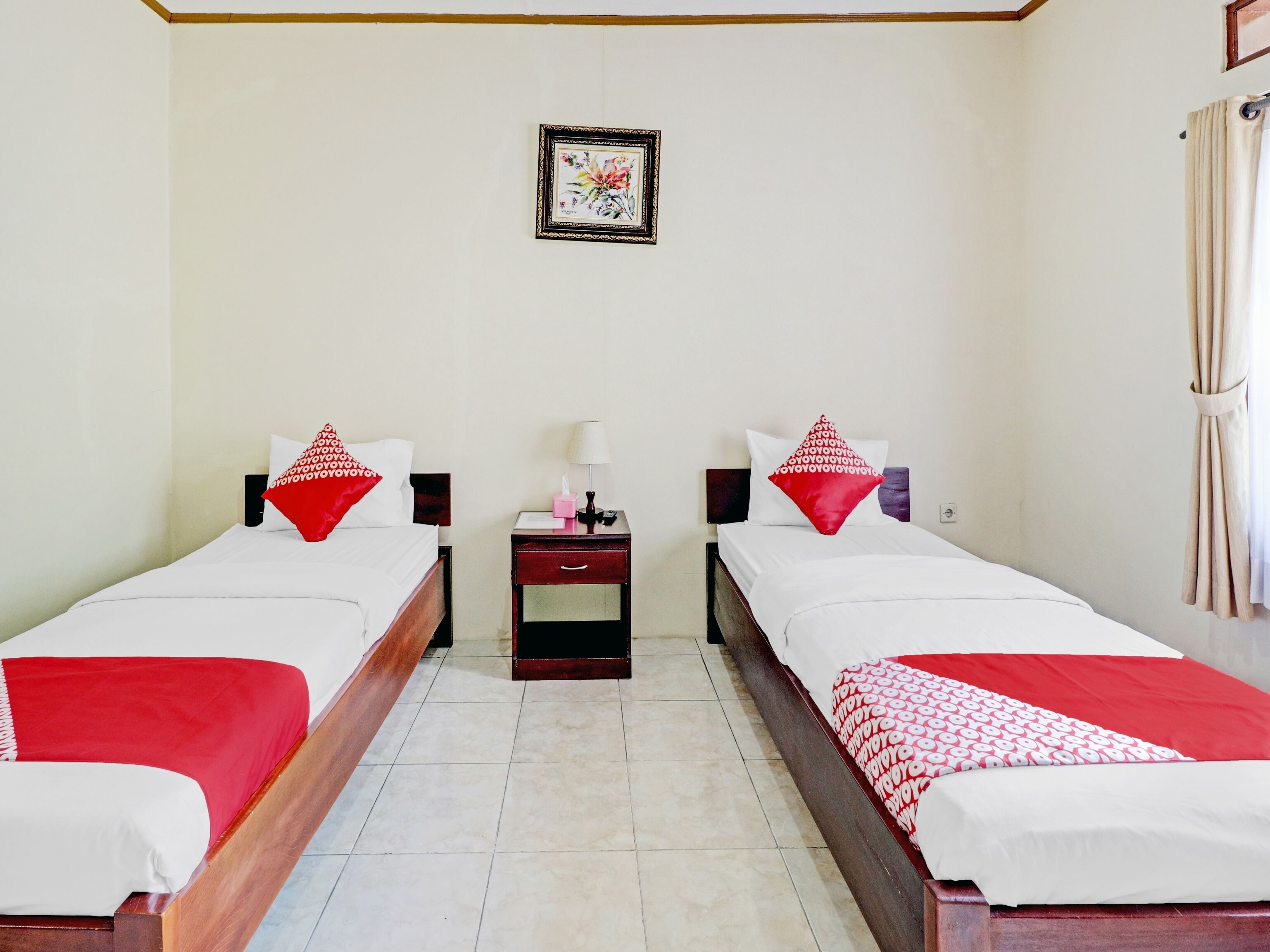 Quarto de hotel com duas camas, cada uma com roupa de cama vermelha e branca, uma pequena mesa de cabeceira com abajur e um quadro na parede.