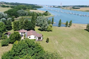 Escape to a serene riverside haven with a two-story house and expansive balcony surrounded by lush greenery and open fields.