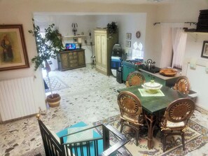 A dining area with a green table and wicker chairs, a kitchen with a stove and cabinets, and a living area with a television and decorative items. - Casa degli Artisti (Nuoro)