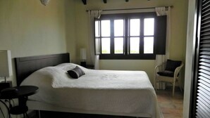 Ein Schlafzimmer mit Bett, Sessel, Tisch und einem fenster mit Vorhängen.