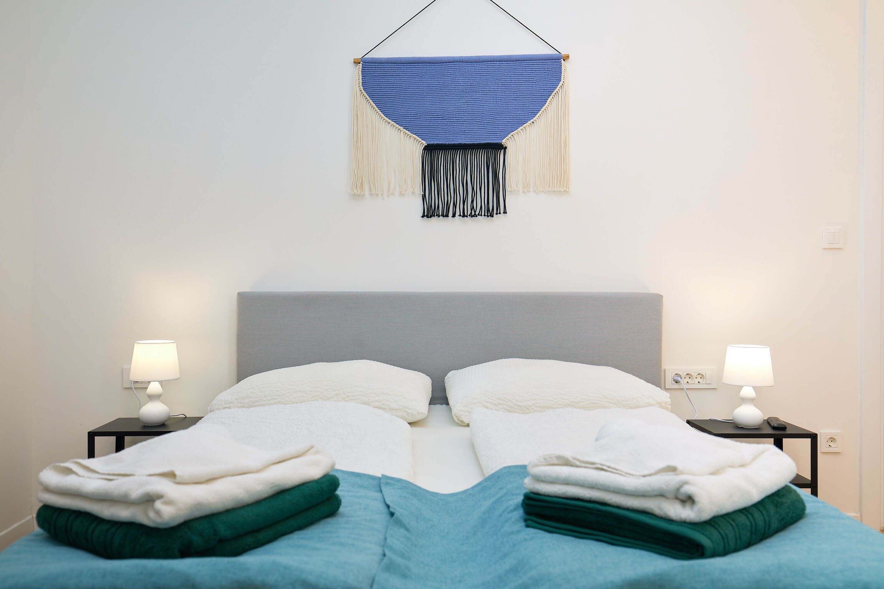 A neatly made bed with folded towels, two bedside lamps, and a wall hanging above.