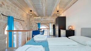 Un dormitorio con pared de piedra, techo de madera, cama con ropa blanca, mesita de noche y vista a un comedor.