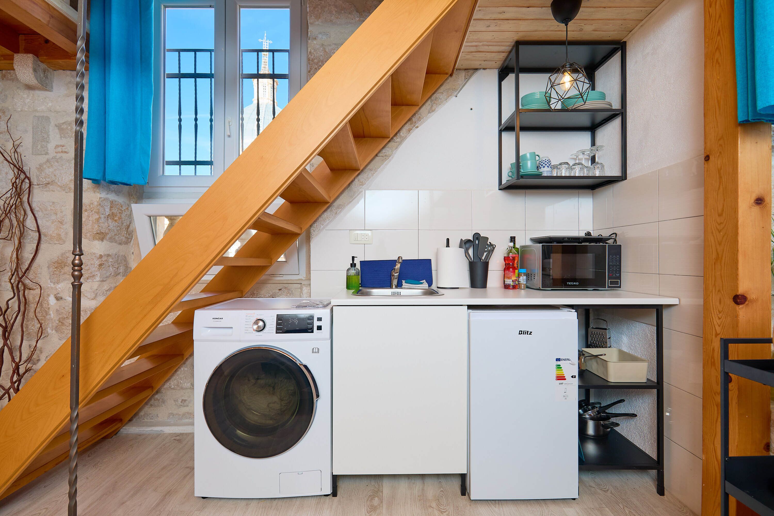 Una cocina compacta con lavadora, refrigerador y microondas. Una escalera de madera conduce a un nivel superior.