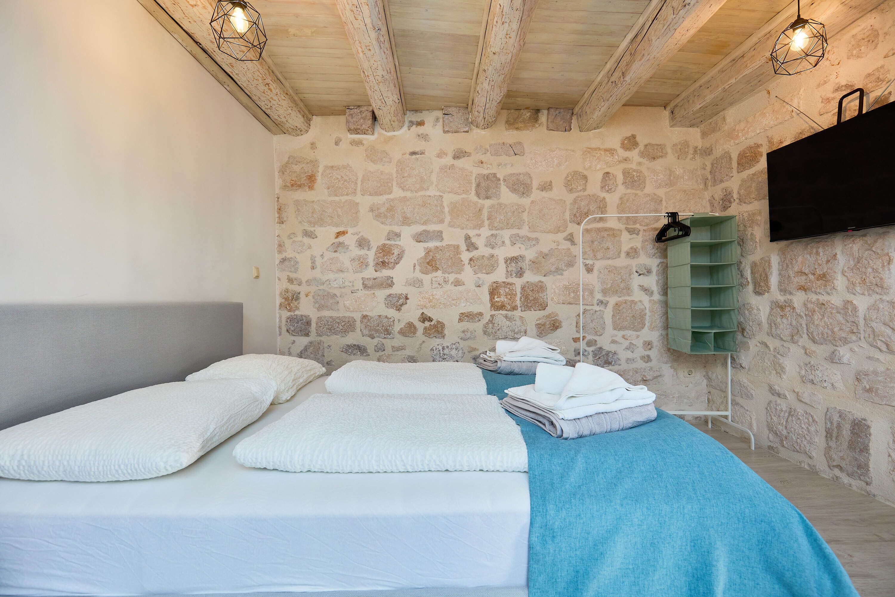 A bedroom with a stone wall, a bed with white linens, a blue blanket, and a TV mounted on the wall.
