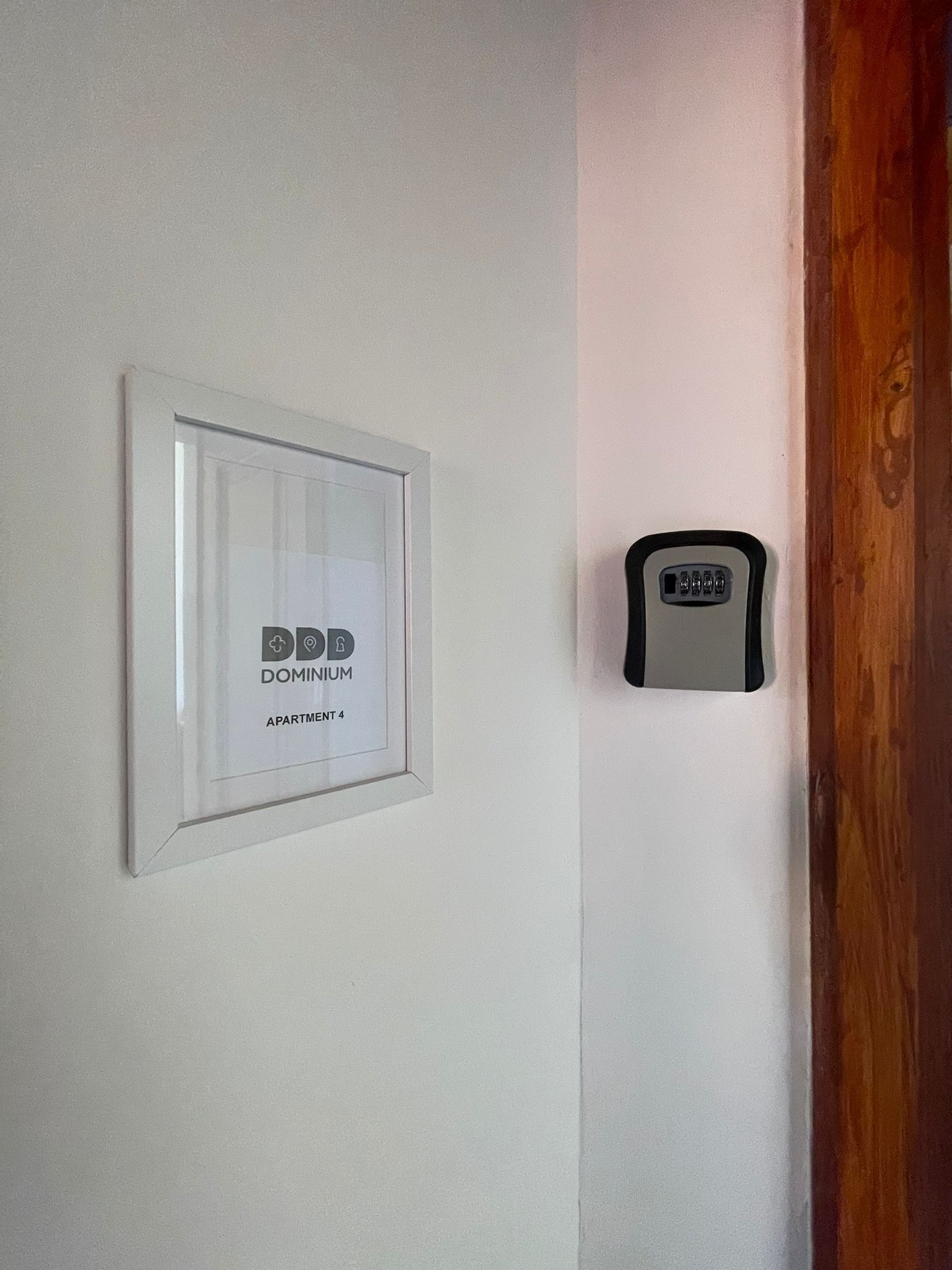Ein gerahmtes Schild mit der Aufschrift 'DOMINIUM APARTMENT 4' an einer weißen Wand, und ein kleines elektronisches Gerät an der angrenzenden Wand.