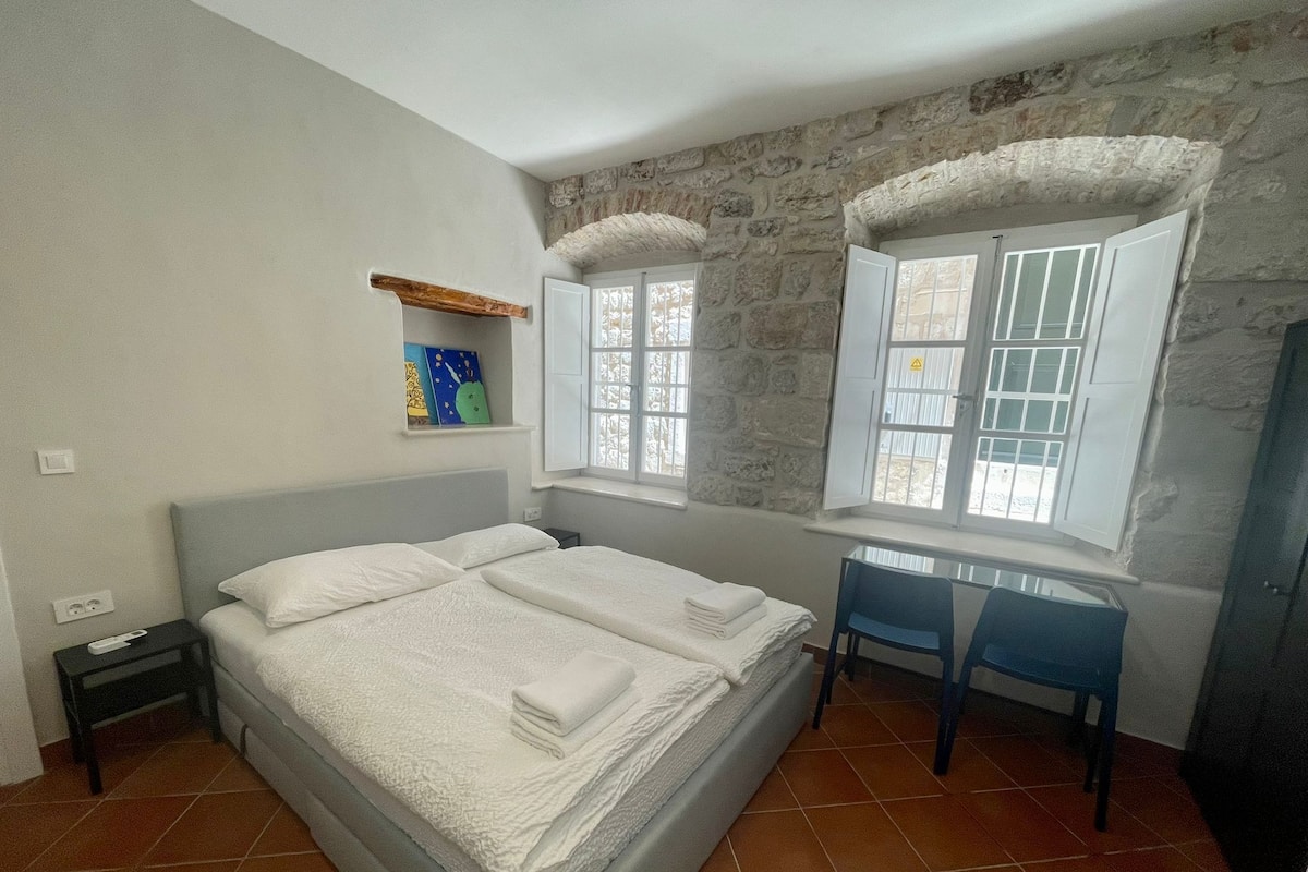 A bedroom with a bed, a desk, and a chair. There is a window with a view of the outside.