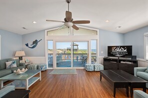 A spacious living room with a large TV, a ceiling fan, and a view of the beach through glass doors.