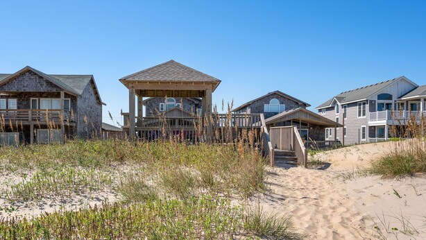 A beachfront property with multiple houses, including a wooden structure with a balcony and a larger house with a covered porch. - Sandy Too-Oceanfront, pool, hot tub, private walkway with gazebo,amazing ocean views! (Nags Head)