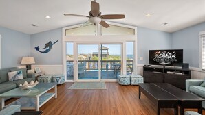 A spacious living room with a large TV, a ceiling fan, and a view of the beach through glass doors. - Sandy Too-Oceanfront, pool, hot tub, private walkway with gazebo,amazing ocean views! (Nags Head)