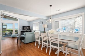 A spacious living room with a dining area, a television, and a view of the beach through large windows.