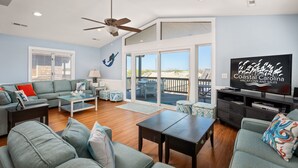 A spacious living room with a large TV, comfortable seating, and a balcony with a view. - Sandy Too-Oceanfront, pool, hot tub, private walkway with gazebo,amazing ocean views! (Nags Head)