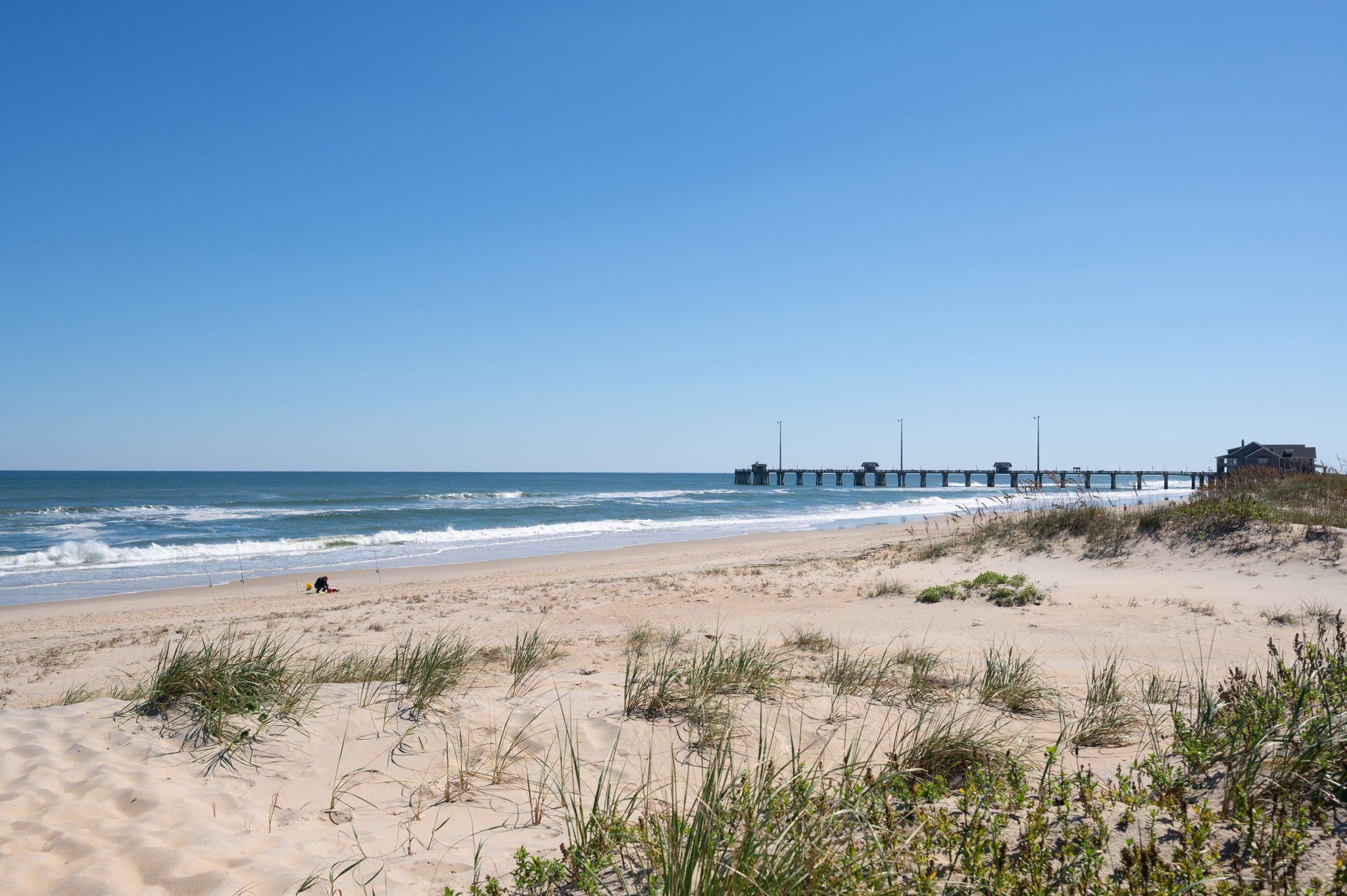 A beach with a pier extending into the ocean, sandy dunes, and a building on the right.