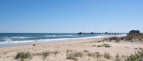A beach with a pier extending into the ocean, sandy dunes, and a building on the right.
