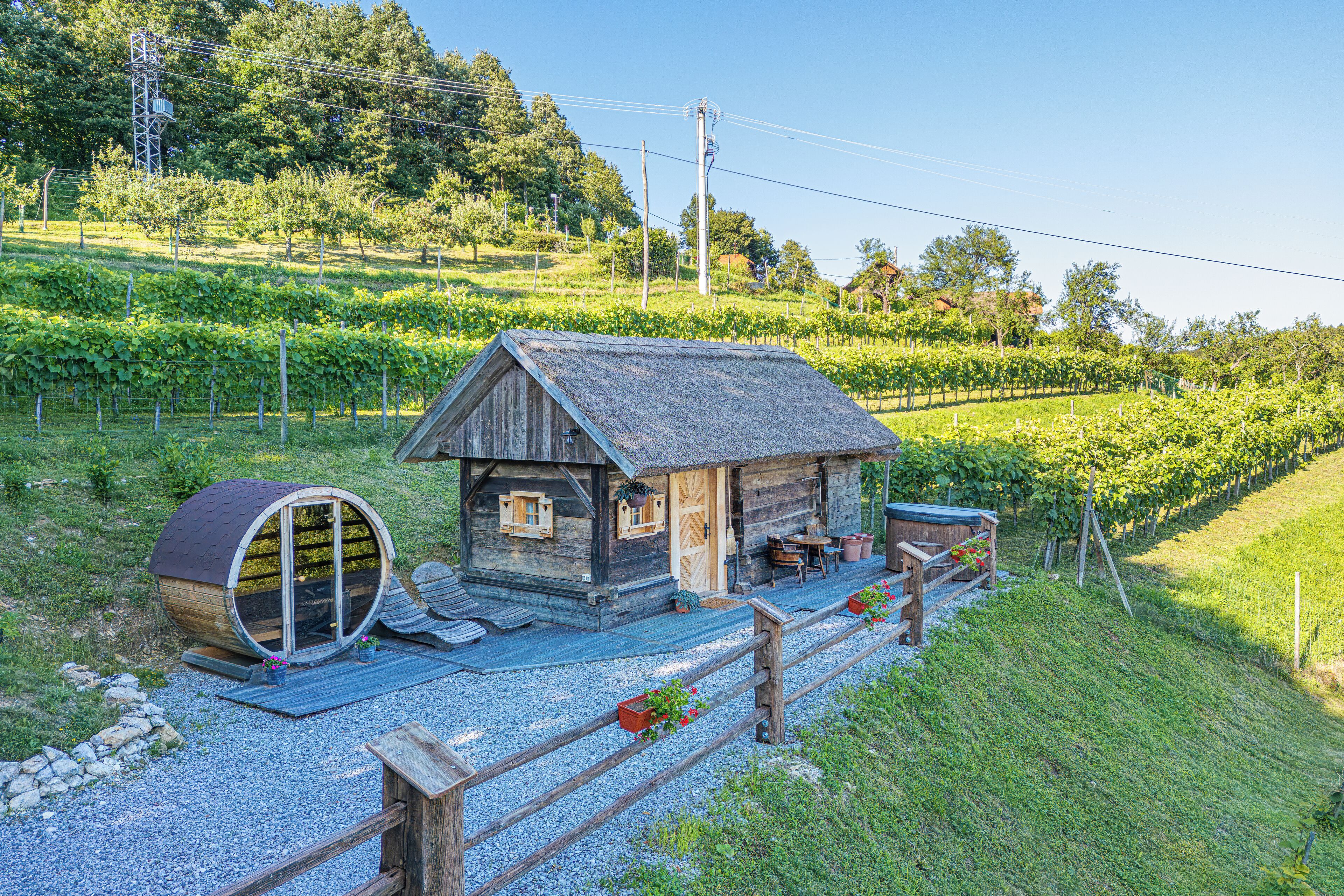 Une cabane rustique avec un toit de chaume, un tonneau en bois et un chemin de gravier menant à l’entrée.
