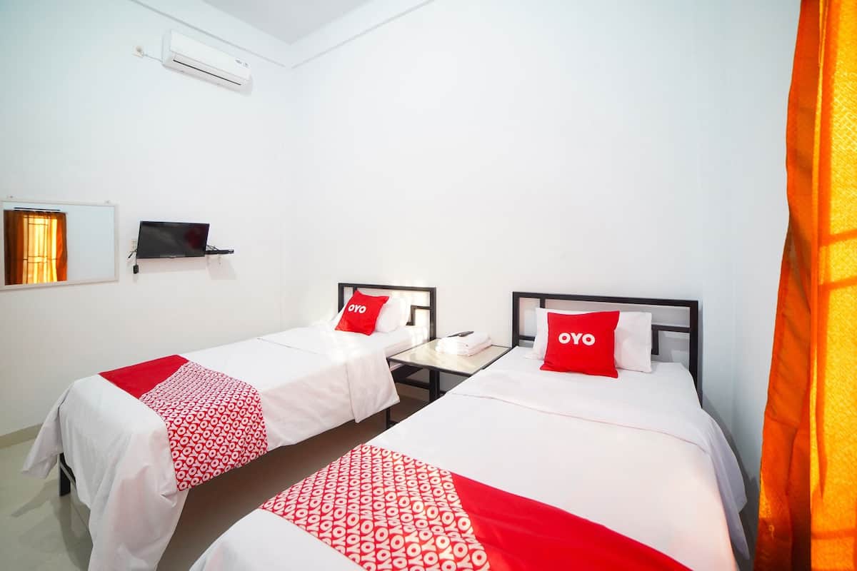 A hotel room with two beds, each with a red and white patterned blanket and a pillow with the word "OYO". There is a small table between the beds, a TV mounted on the wall, and an air conditioning unit above the beds.