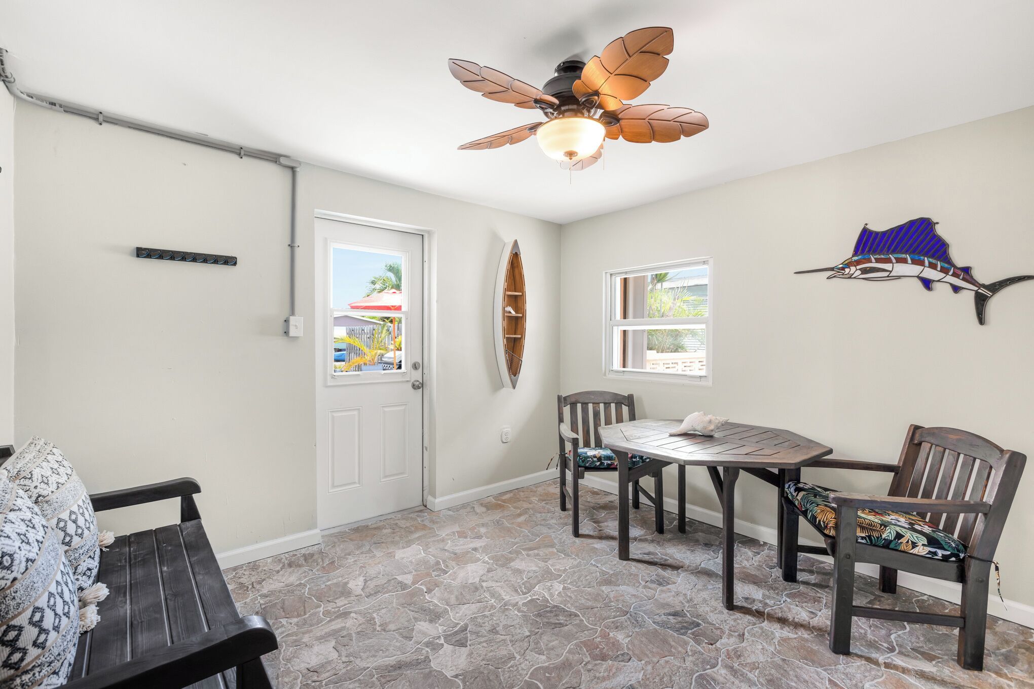 Um quarto com soalho de pedra, um ventilador de teto, uma mesa com cadeiras, uma porta e uma decoração de peixe fixada na parede.