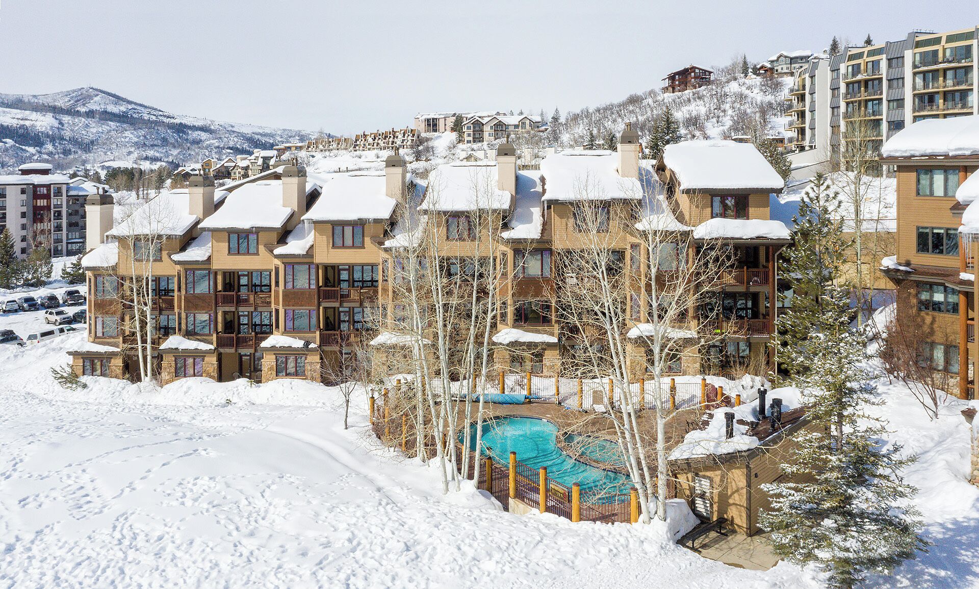 A snow-covered resort with multi-story buildings, a swimming pool, and a mountainous backdrop.