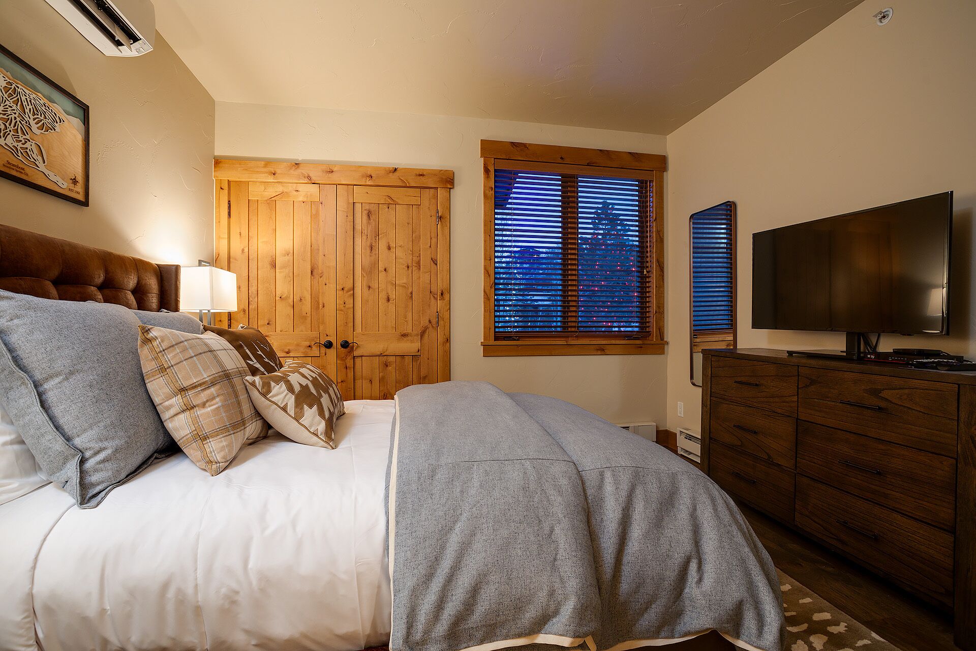 Une chambre à coucher comprenant un lit, une commode, un téléviseur et une fenêtre avec des stores.